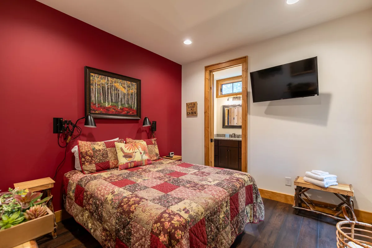 This bedroom scene highlights a plush bed with a patchwork quilt, a wall-mounted TV, a nightstand with plants, and a framed landscape painting. The setup exudes a warm and welcoming vibe and includes amenities like the TV, echoing the master suite's features.