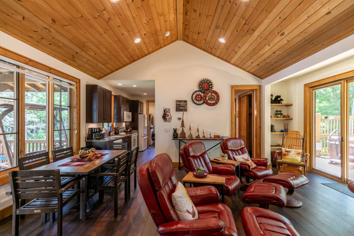 A view of the dining area with red reclining chairs, a wooden dining table, and large windows letting in serene wooded views. Decorative wall art adds splashes of cheerful color, enhancing the vibrant and inviting feel of the space.