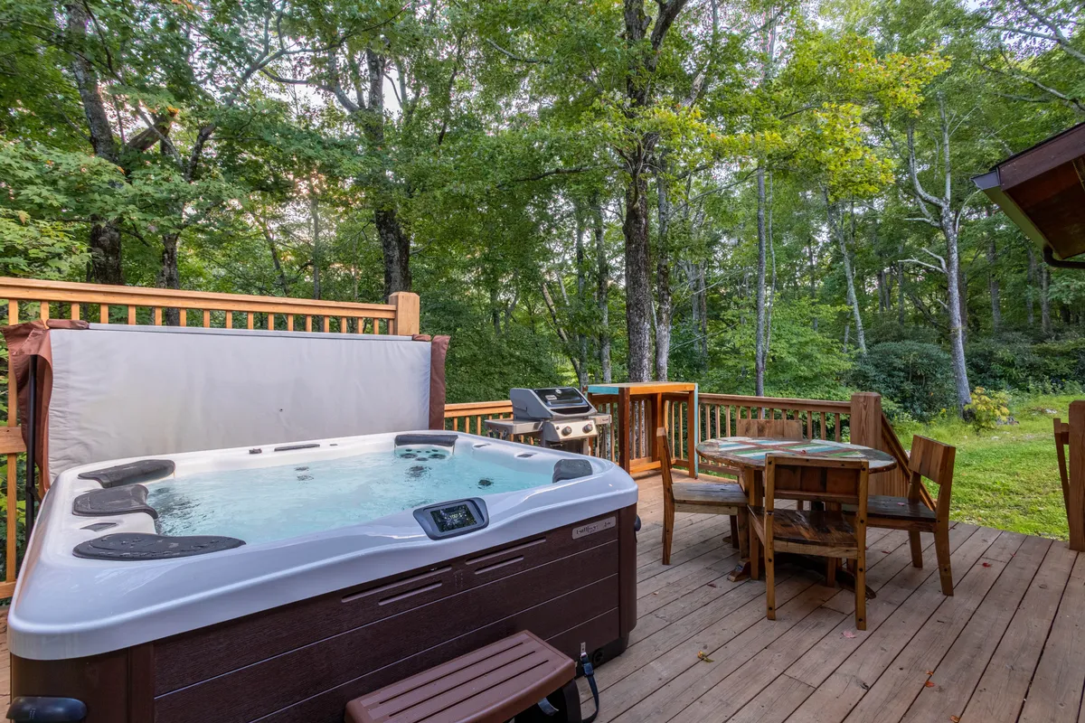 A spacious wooden deck features a luxurious hot tub, dining table, grill, and is surrounded by tall trees. The setup is ideal for relaxing after a day outdoors, enjoying nature, and social gatherings.