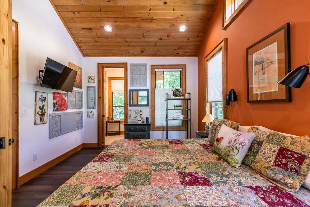 This image features a cozy bedroom with a patchwork quilt on the bed, a wooden ceiling, decorative wall art, and a nightstand with a lamp. The warm lighting and soft textures evoke comfort and relaxation—key aspects of a private retreat. The visible window hints at natural views and the peaceful hush surrounding the cottage.