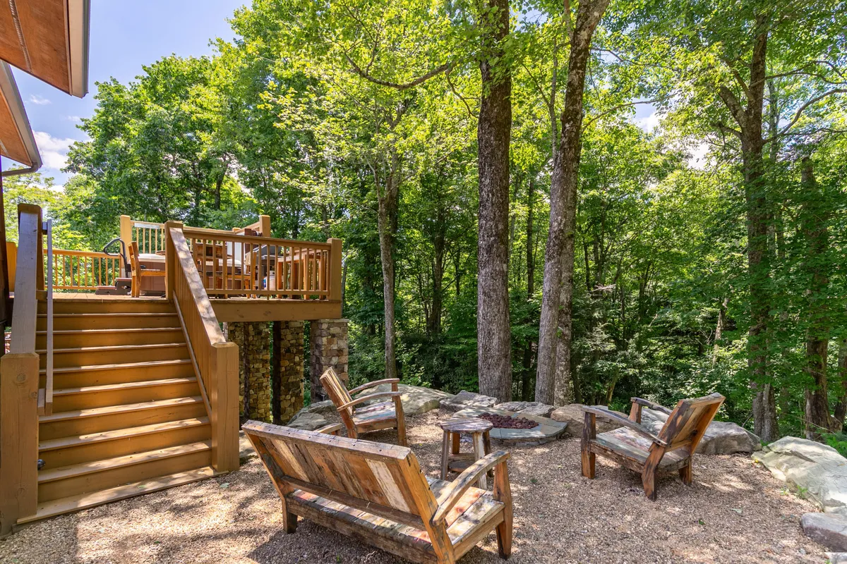 A cozy wooden deck with a fire pit and comfortable chairs is set amidst a forested area. The scene suggests relaxation, storytelling around the fire, and enjoying the sounds of nature, with ample space for groups to gather.
