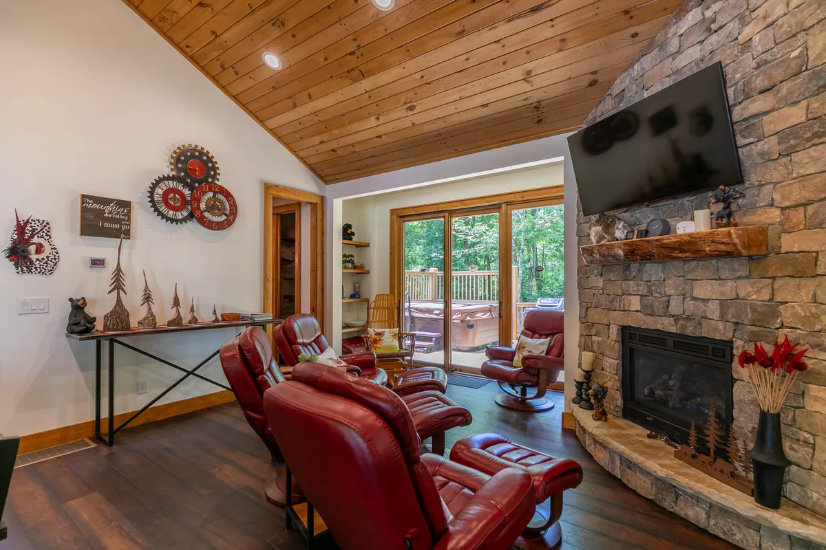 A welcoming living room with a stone fireplace as the focal point, flanked by reclining chairs. Large glass doors open to a deck, creating a visual transition between the cozy interior and the outdoors. Sunlight streams in, hinting at the beauty of the surrounding area and inviting guests to explore beyond the home.