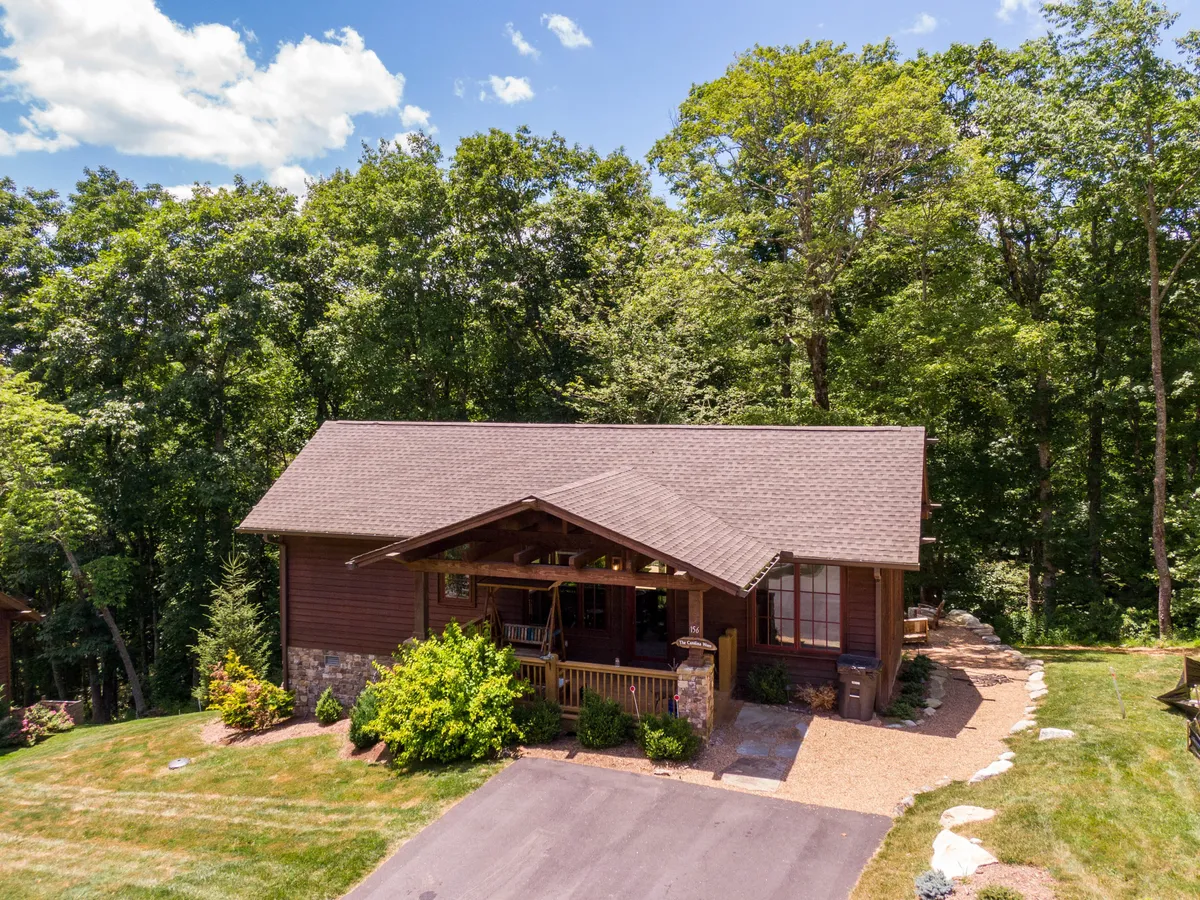 The image showcases the charming exterior of Carolina Wren Cottage, with a welcoming wooden porch furnished with classic rocking chairs, surrounded by a beautifully landscaped garden and natural stone accents on the house. The tranquil scene hints at sunset views and the serenity of the mountain setting.