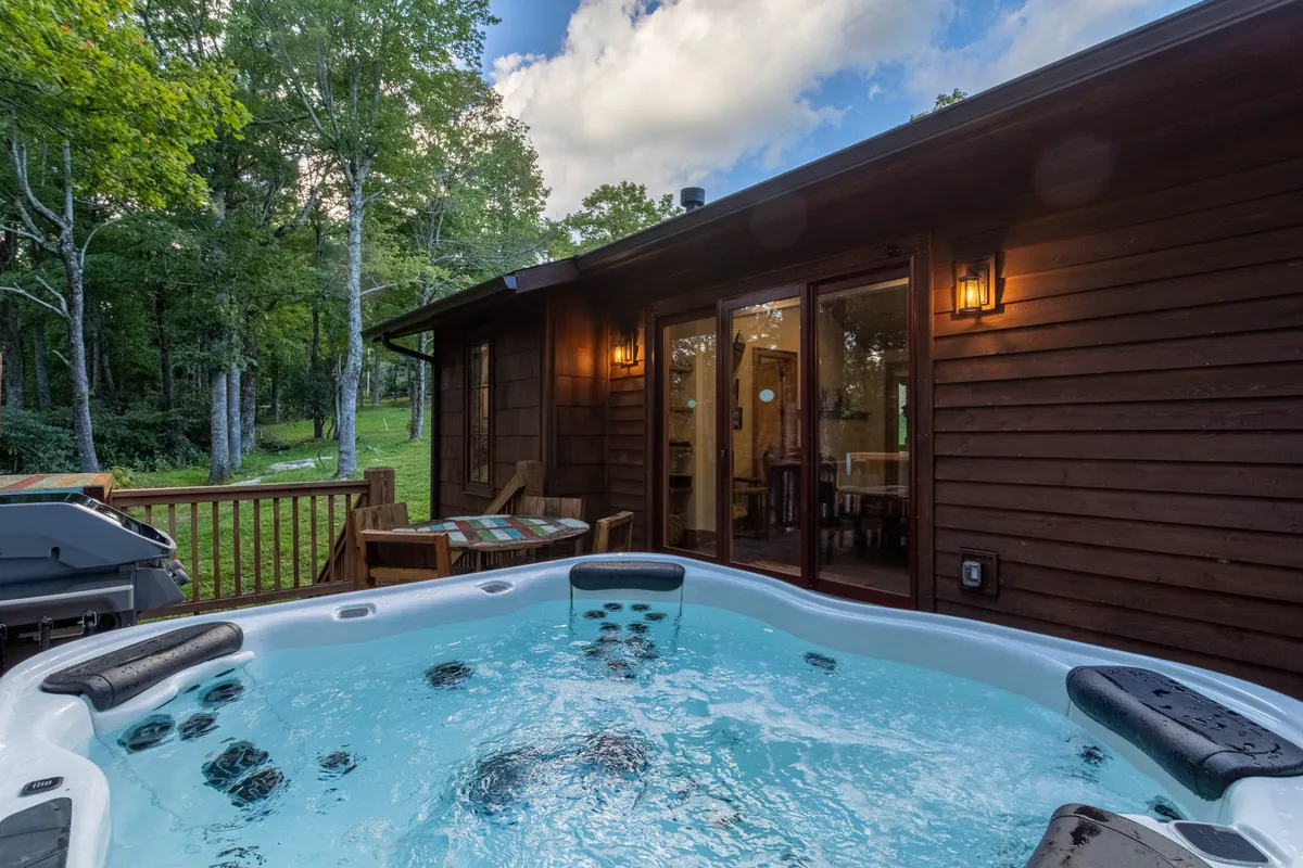 A tranquil view of the hot tub set on the wooden deck, framed by lush trees and adjacent to glass doors leading inside. The image evokes serenity, perfect for moments of soaking while immersed in nature, and hints at the seamless indoor-outdoor flow.